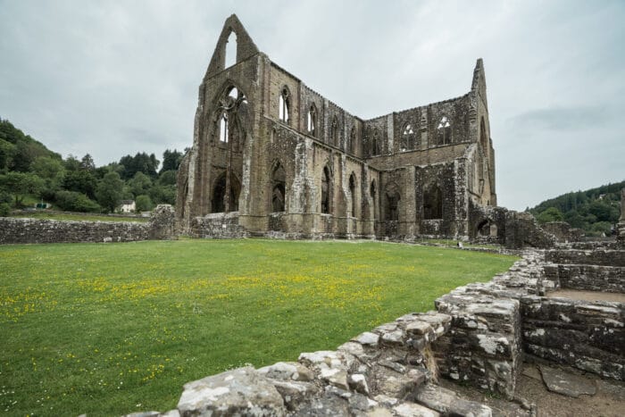 The image shows the stone ruins of Tintern Abbey, a large Gothic abbey with tall arched windows, surrounded by green grass and low stone walls under a cloudy sky. Trees and hills can be seen in the background.