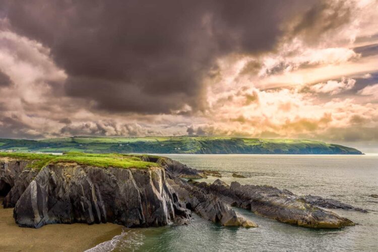 Dramatic clouds and sunlight illuminate the rugged Welsh coastline in Wales with steep cliffs, green grassy tops, jagged rocks, and calm sea water along a scenic shore.