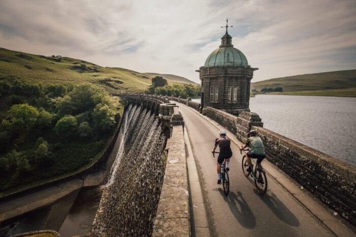 Two cyclists ride along a stone dam with a domed tower, part of one of the scenic cycling routes in Wales, surrounded by green hills and water under a partly cloudy sky.