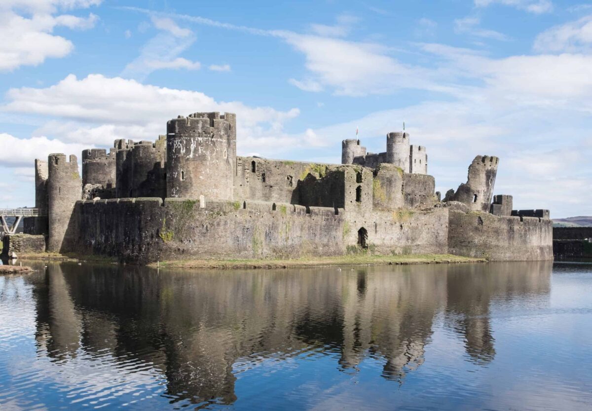 A bright daytime view of Caerphilly Castle surrounded by a serene defensive moat with its famous leaning tower visible on the right, offering fantastic heritage and family days out in Wales.