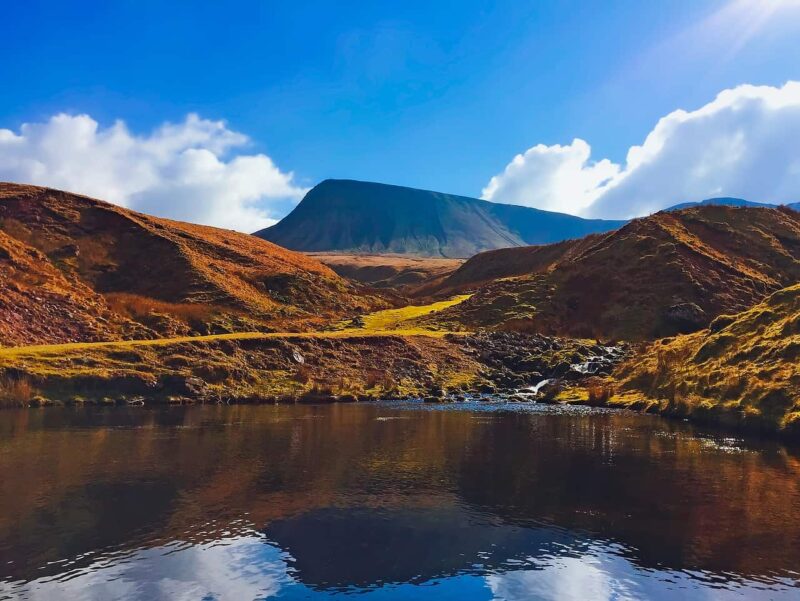 A clear blue sky with scattered clouds over a mountain and rolling brown hills, reflected in a calm stream or pond in the foreground. Bright sunlight highlights the landscape. A perfect hiking in Wales location
