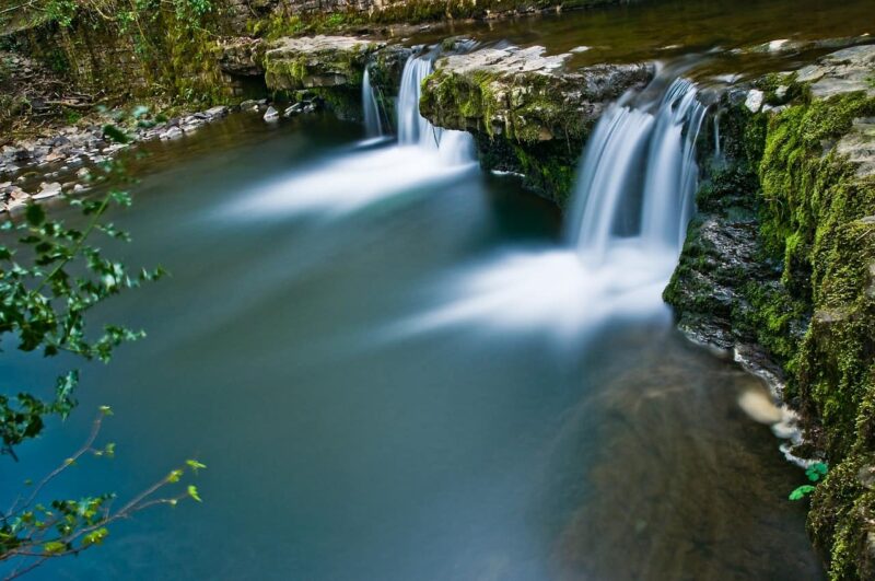A small waterfall cascades over moss-covered rocks into a calm, clear pool surrounded by greenery, captured with a long exposure for a silky water effect on a picturesque waterfall walk in Wales.
