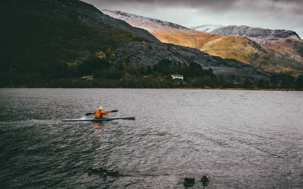 A person in an orange jacket enjoys sea kayaking in Wales on a calm lake, with ducks swimming nearby. Hills and mountains rise in the background, partially lit by sunlight breaking through cloudy skies.