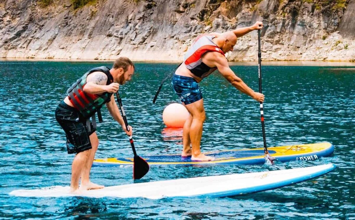 Two men wearing life jackets paddle standing on separate boards, paddleboarding in Wales on clear blue water near a rocky shoreline. One man leans forward as he paddles, while a large buoy floats in the background.