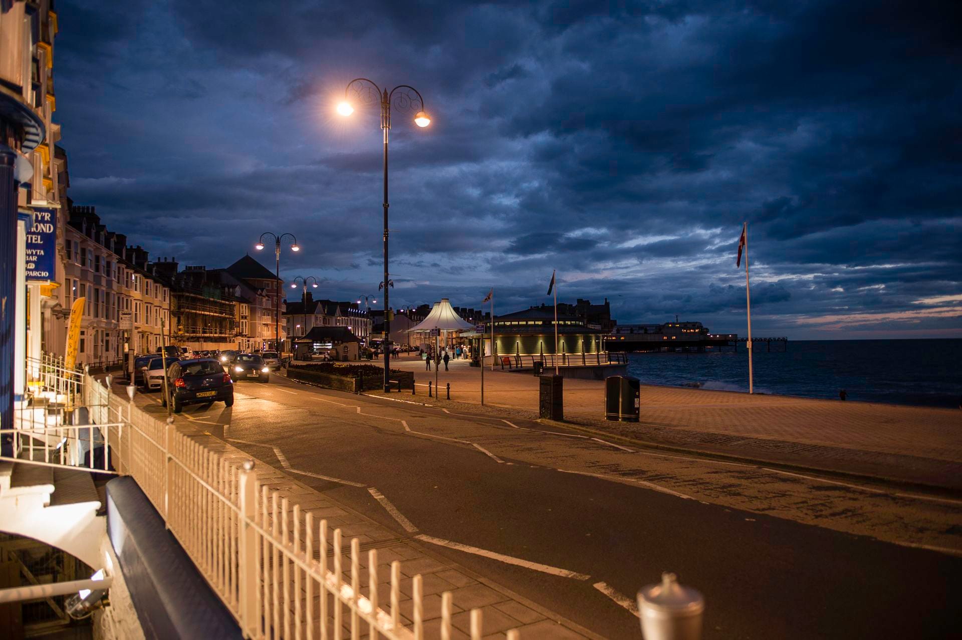 The seafront of Aberystwyth at twilight, with the curved Victorian promenade and pastel-coloured terraced houses illuminated against the evening sky, and the lights of Cardigan Bay reflecting in the calm waters below.