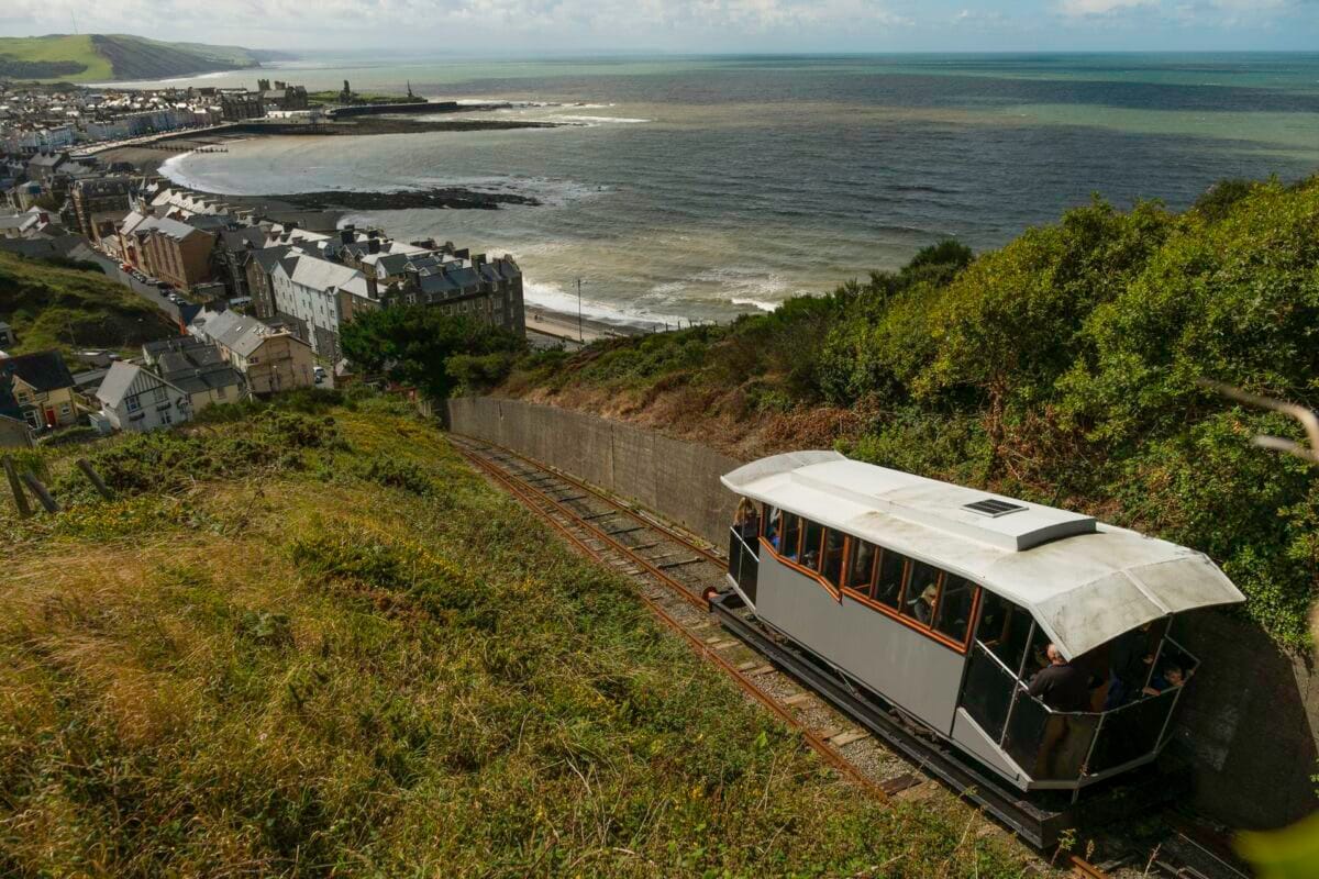 A vintage funicular cliff railway carriage climbing a steep, grassy hill, looking down over the sweeping bay, dark Irish Sea, and curved Victorian seafront of Aberystwyth.