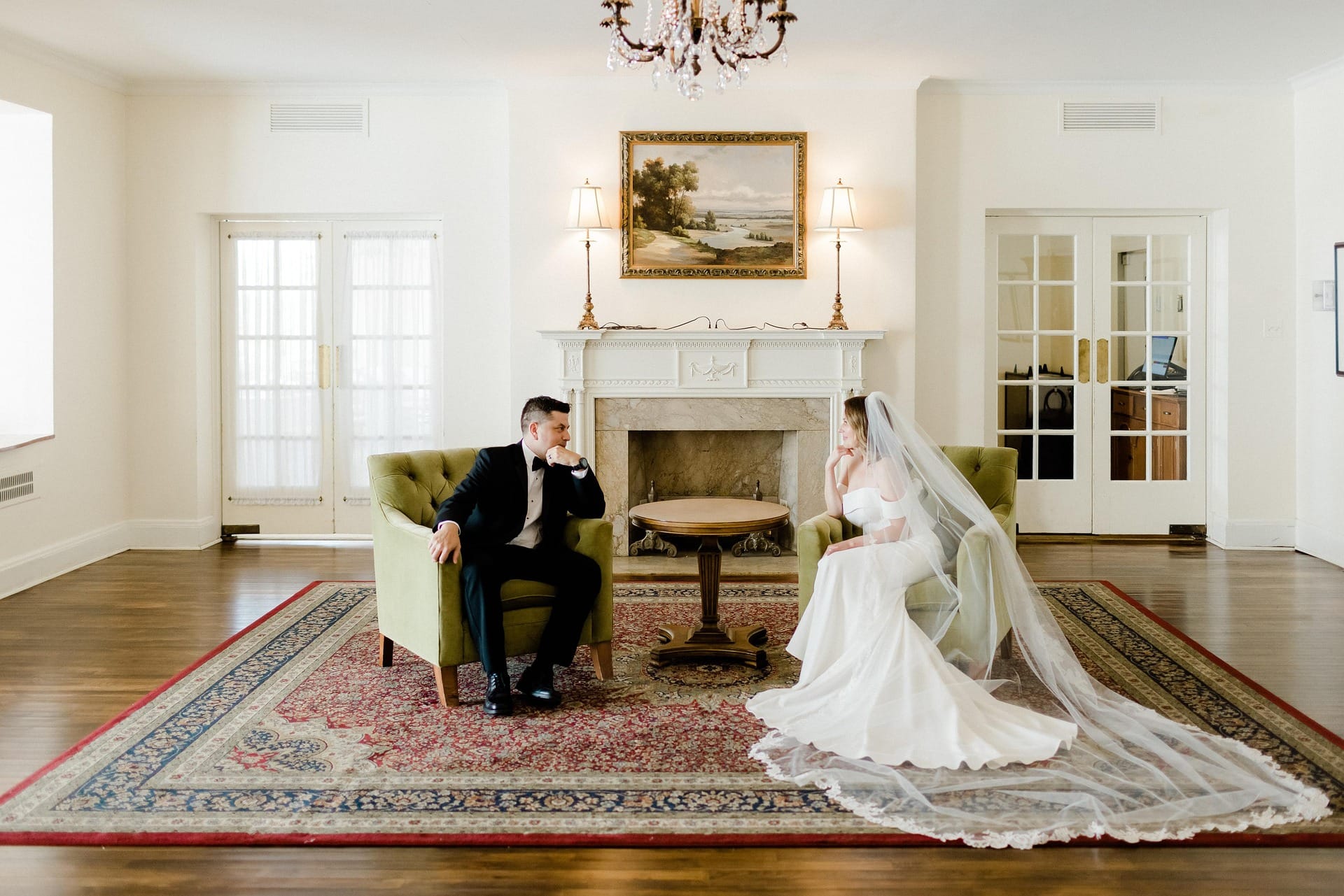 A bride and groom sitting across from each other in pale green armchairs in a spacious, elegant room. The bride is wearing a white gown with a long, flowing veil trailing across a large patterned area rug, and the groom is in a classic black tuxedo. They are facing each other in front of a white fireplace mantel and a landscape painting.