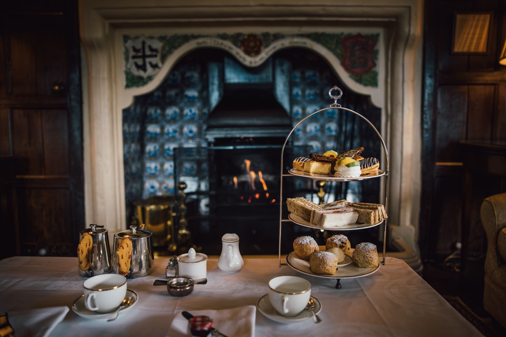 A traditional three-tier afternoon tea stand filled with sandwiches, scones, and pastries, set on a white linen table alongside fine china teacups and silver teapots. In the background, a warm fire burns in a grand, historic stone fireplace decorated with blue and white tiles inside a dark wood-panelled room.