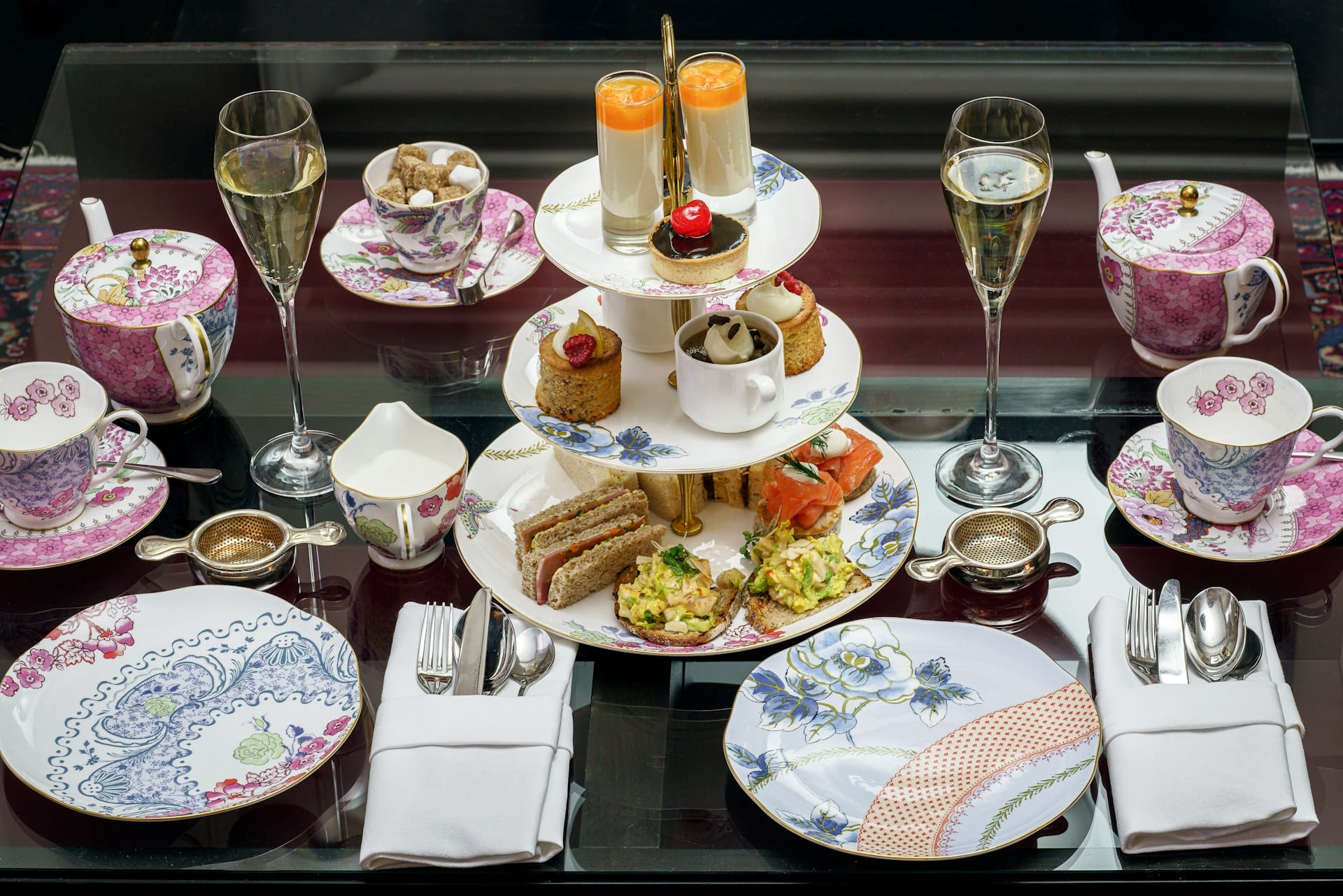 A luxurious afternoon tea set on a dark reflective table. In the center is a three-tier stand holding finger sandwiches, small cakes, and delicate desserts. Two glasses of Champagne flank the stand, surrounded by ornate pink and blue floral teacups, saucers, patterned side plates, and a matching teapot.