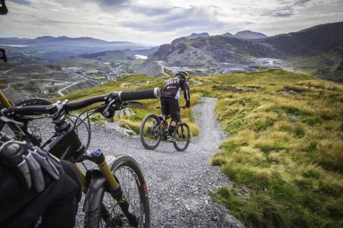 2 mountain bikers going down a gravel trail in Snowdonia with the mountains in the background on a cloudy day