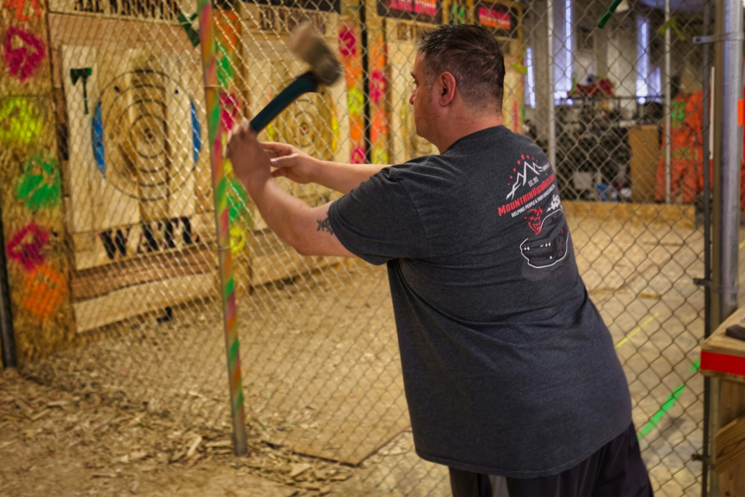 A man viewed from behind swings a small axe with both hands, mid-throw toward a wooden bullseye target inside a protective chain-link throwing lane at an indoor urban axe-throwing venue