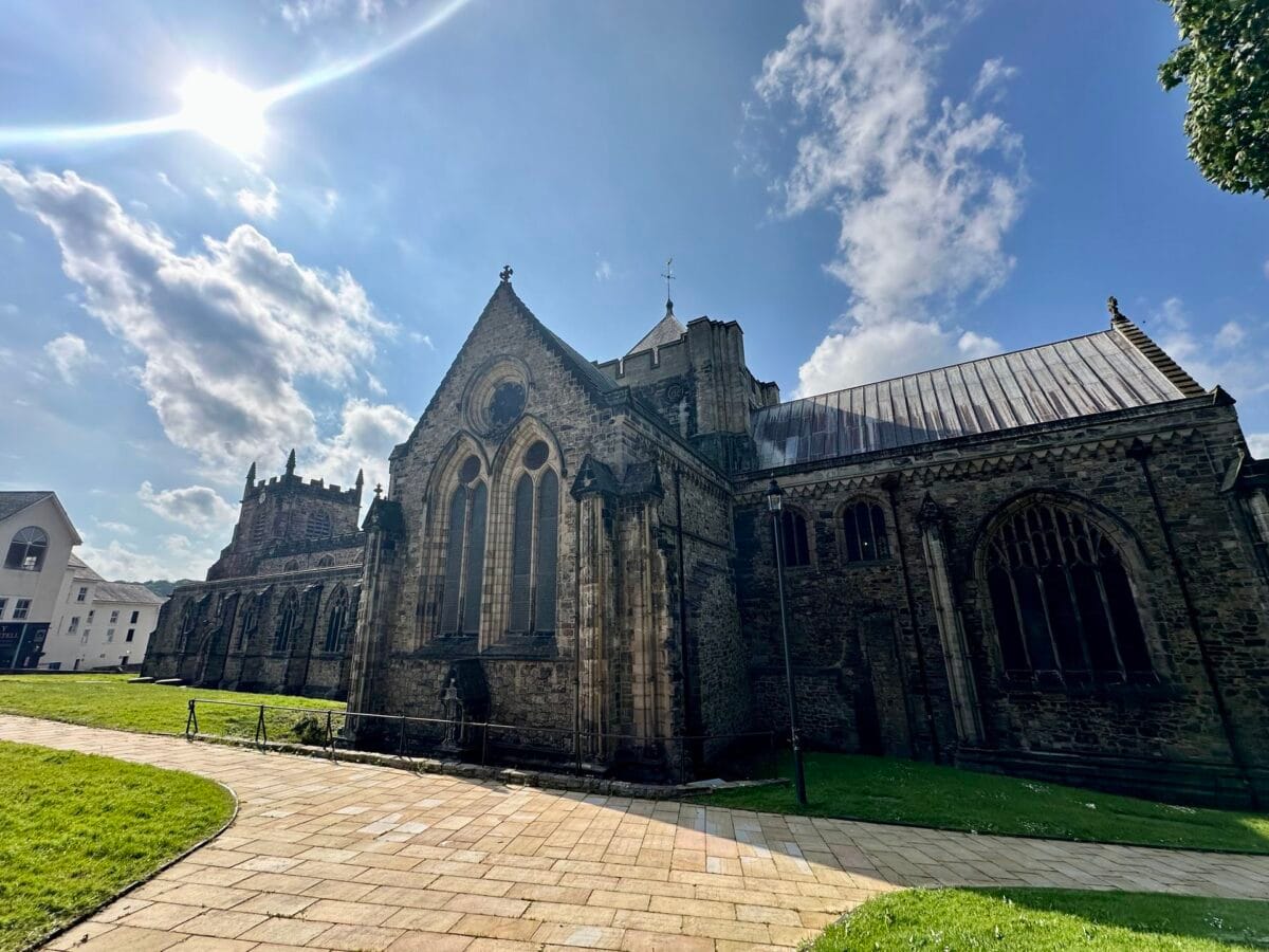 The historic stone exterior of Bangor Cathedral featuring gothic arched windows and a paved walkway, set against a bright sunny sky