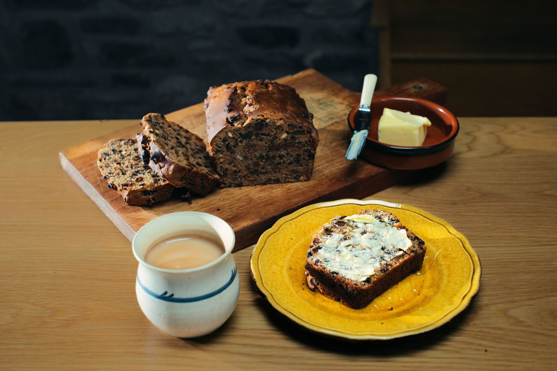 A sliced loaf of traditional Welsh bara brith on a rustic wooden board, alongside a thick buttered slice served on a yellow plate next to a ceramic mug of hot Welsh tea.
