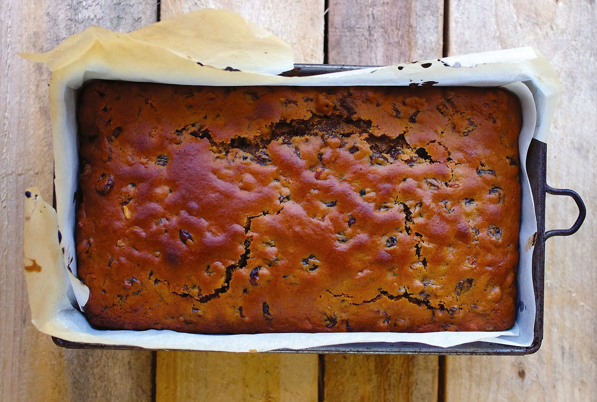 An overhead view of a freshly baked loaf of traditional Welsh bara brith fruit cake studded with dried raisins, cooling inside a parchment-lined metal baking tin.