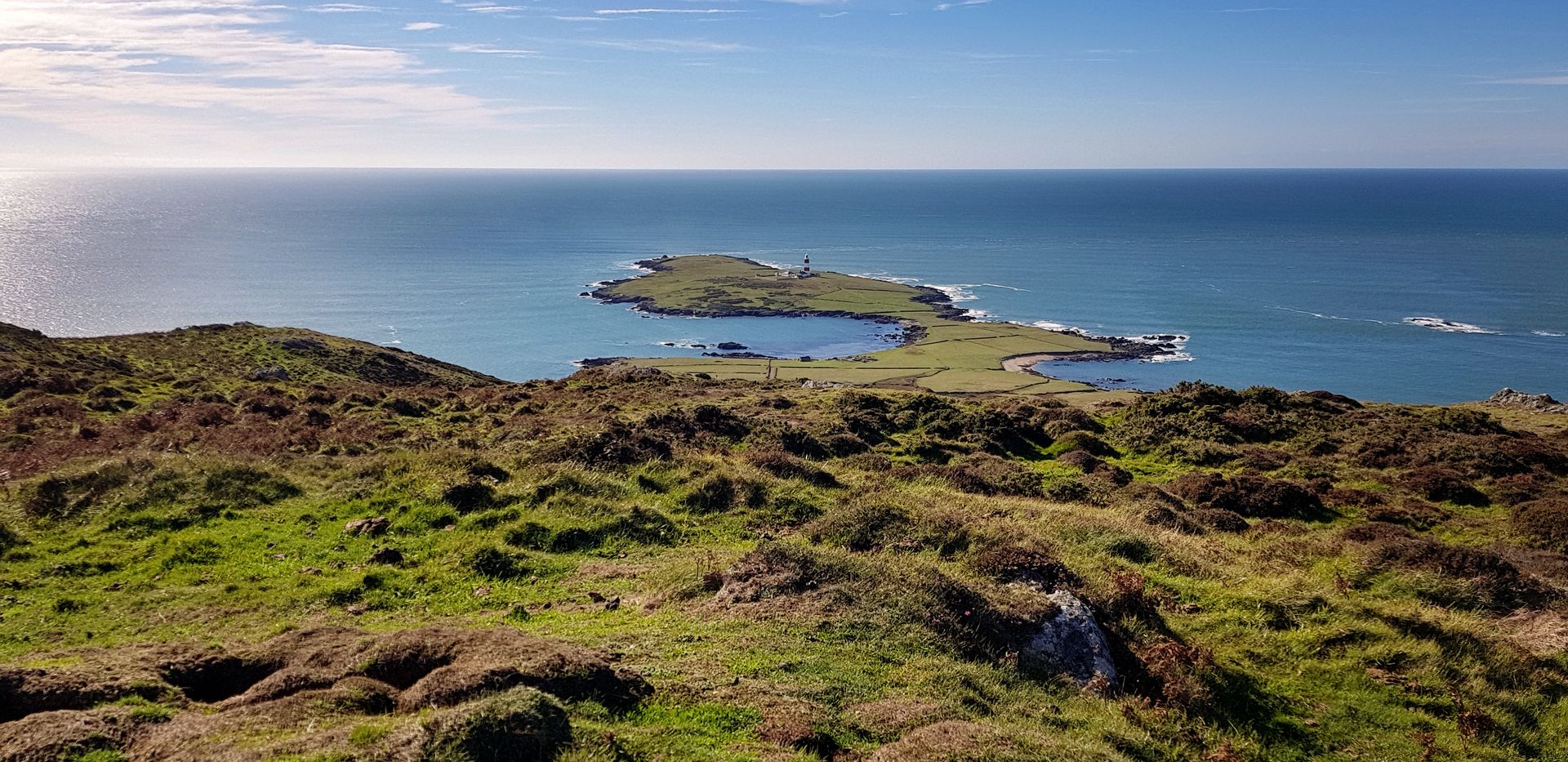 A sweeping, high-angle view from the hills of Bardsey Island. Rugged green and brown moorland in the foreground gives way to a narrow peninsula stretching into a vast, bright blue sea. A white lighthouse stands at the very edge of the land under a clear, sunlit sky.