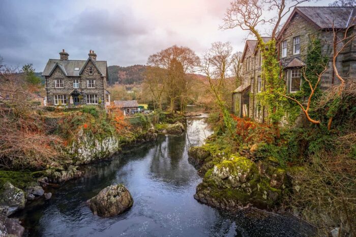 A tranquil river flows between two stone houses in Betws Y Coed, surrounded by trees and lush vegetation, with rocky banks and hills in the background under a cloudy sky.