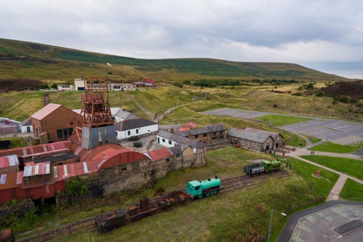 Aerial view of the Big Pit Museum, showcasing historic mining buildings, rusted structures, railway tracks, and a green railway carriage, all set amid grassy hills under an overcast sky.