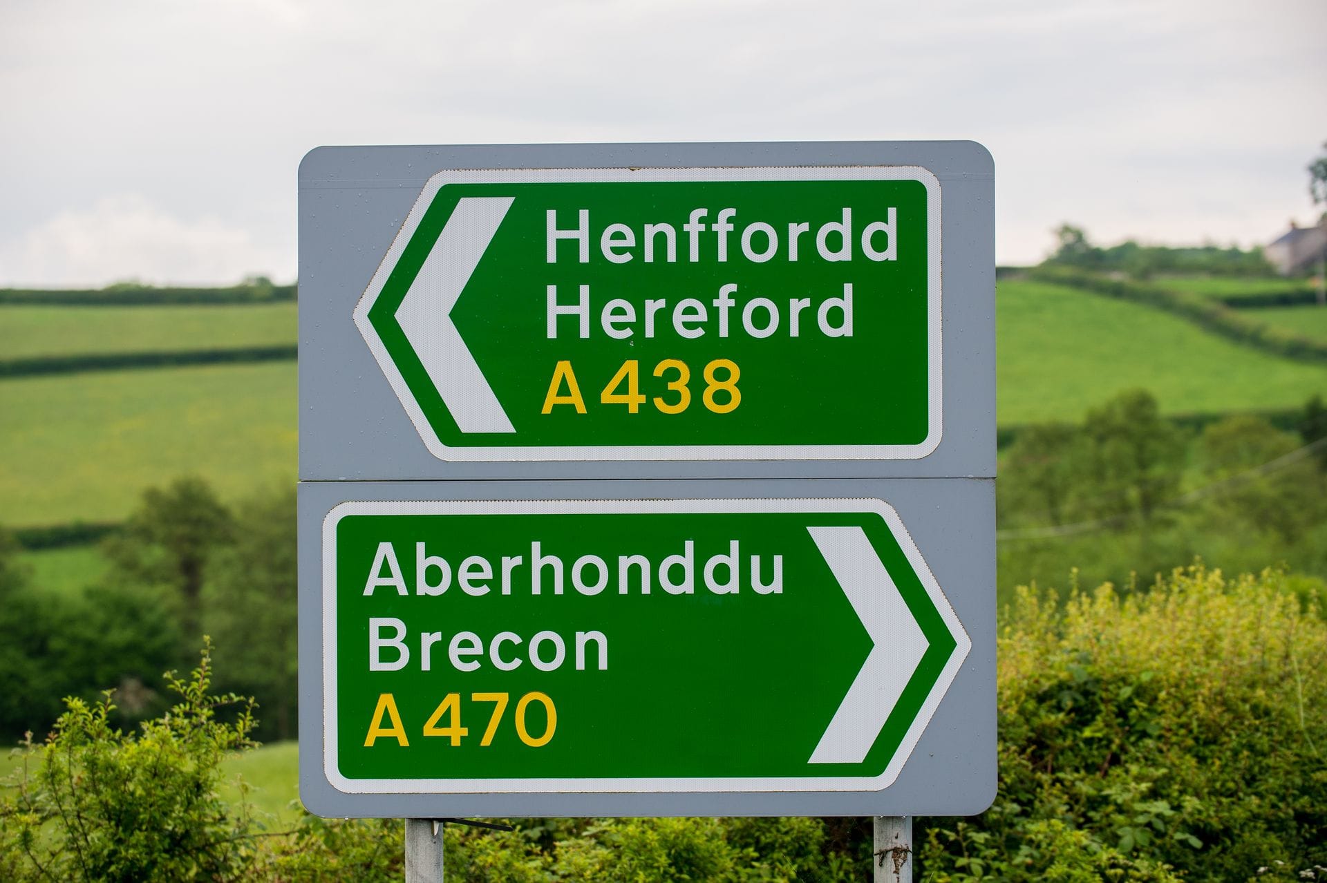 A close-up photograph of a standard green and white directional road sign in Wales, showing bilingual place names in Welsh and English with mileage markers, positioned beside a rural roadside.