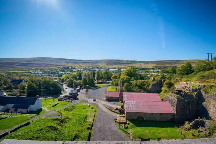 A bright, high-angle view overlooking the historic Blaenavon Ironworks in the South Wales Valleys on a sunny day. The heritage site features a collection of preserved stone industrial buildings, a tall chimney, and a structure with a prominent red roof, all surrounded by vibrant green lawns and set against a backdrop of rolling hills under a clear blue sky.
