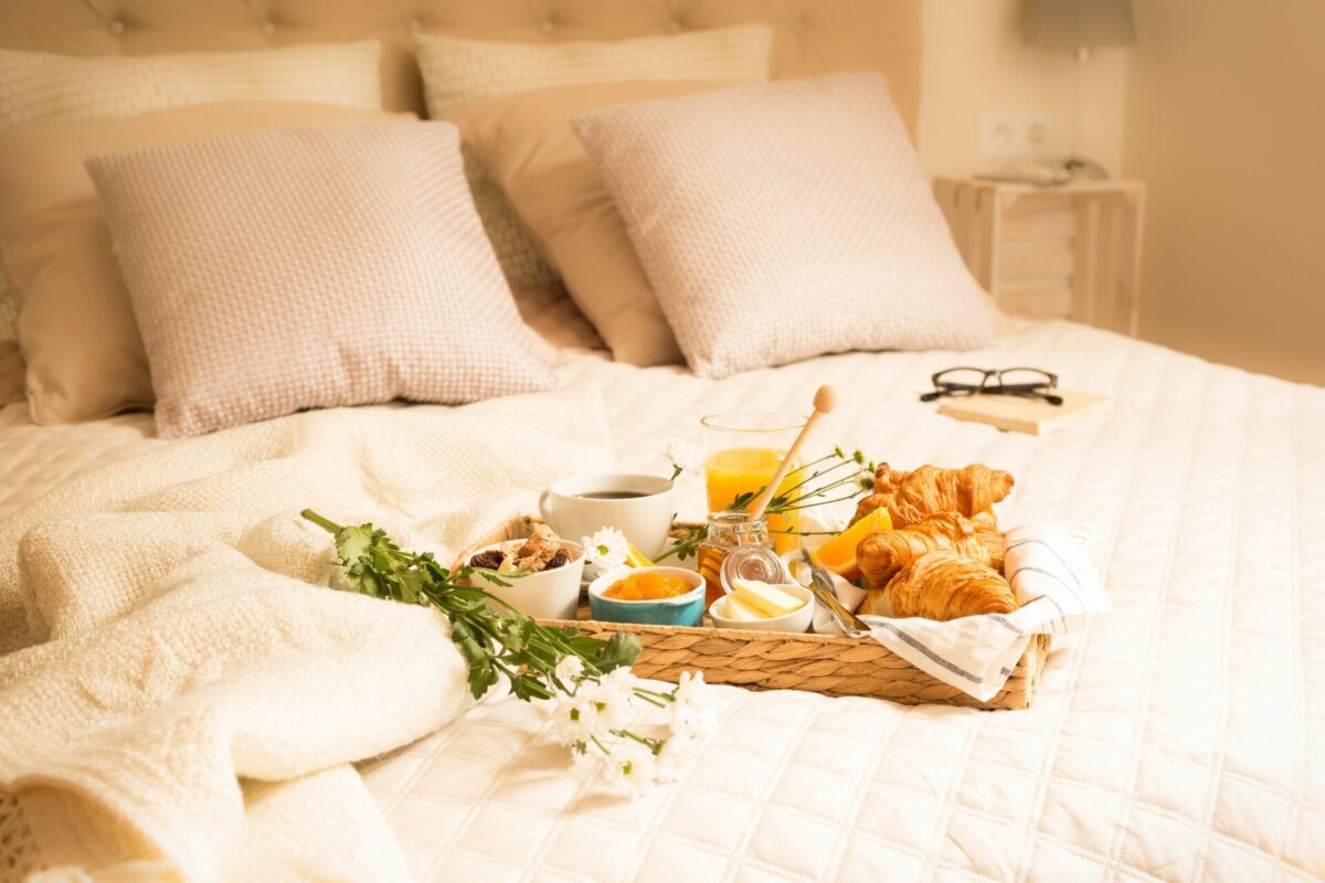 A luxurious romantic breakfast in bed featuring fresh croissants, orange juice, and coffee on a woven tray resting on a cozy hotel bed with white linens in a Swansea Bay hotel