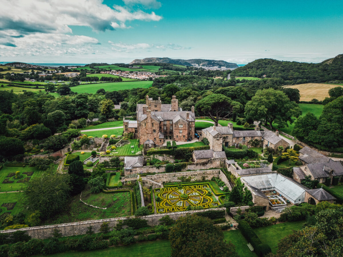 Aerial view of Bodysgallen Hall spa hotel in Wales, an old stone mansion surrounded by lush gardens and green fields, with a patterned formal garden and glasshouse; rolling hills and a village appear in the distant background under a blue sky. One of the best hotels in Wales