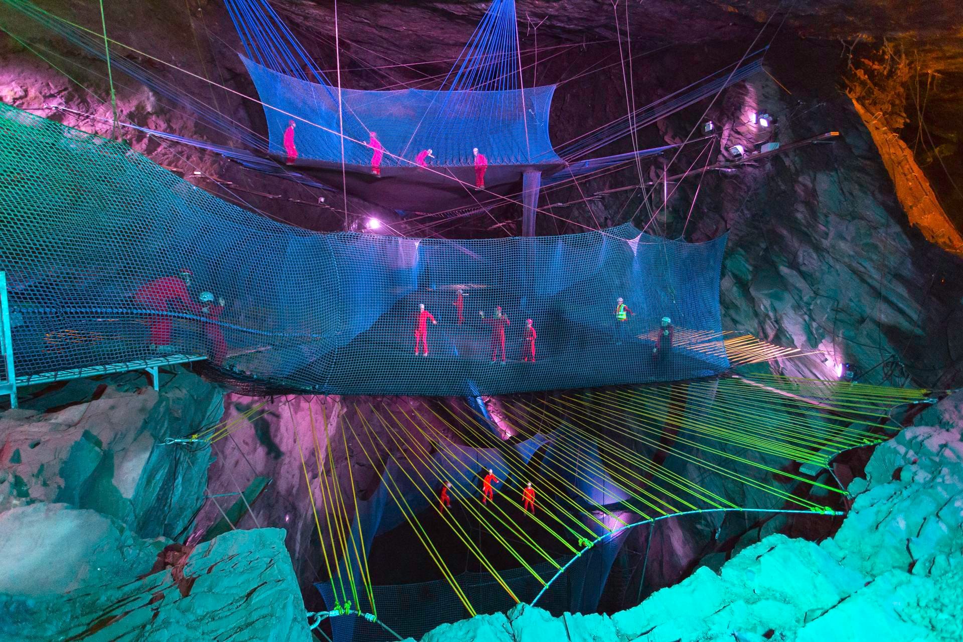 A massive underground slate cavern illuminated with vibrant neon pink and teal lights. Multiple tiers of large blue trampoline nets are suspended high in mid-air by cables, with small figures wearing bright red jumpsuits bouncing and walking across the different levels.