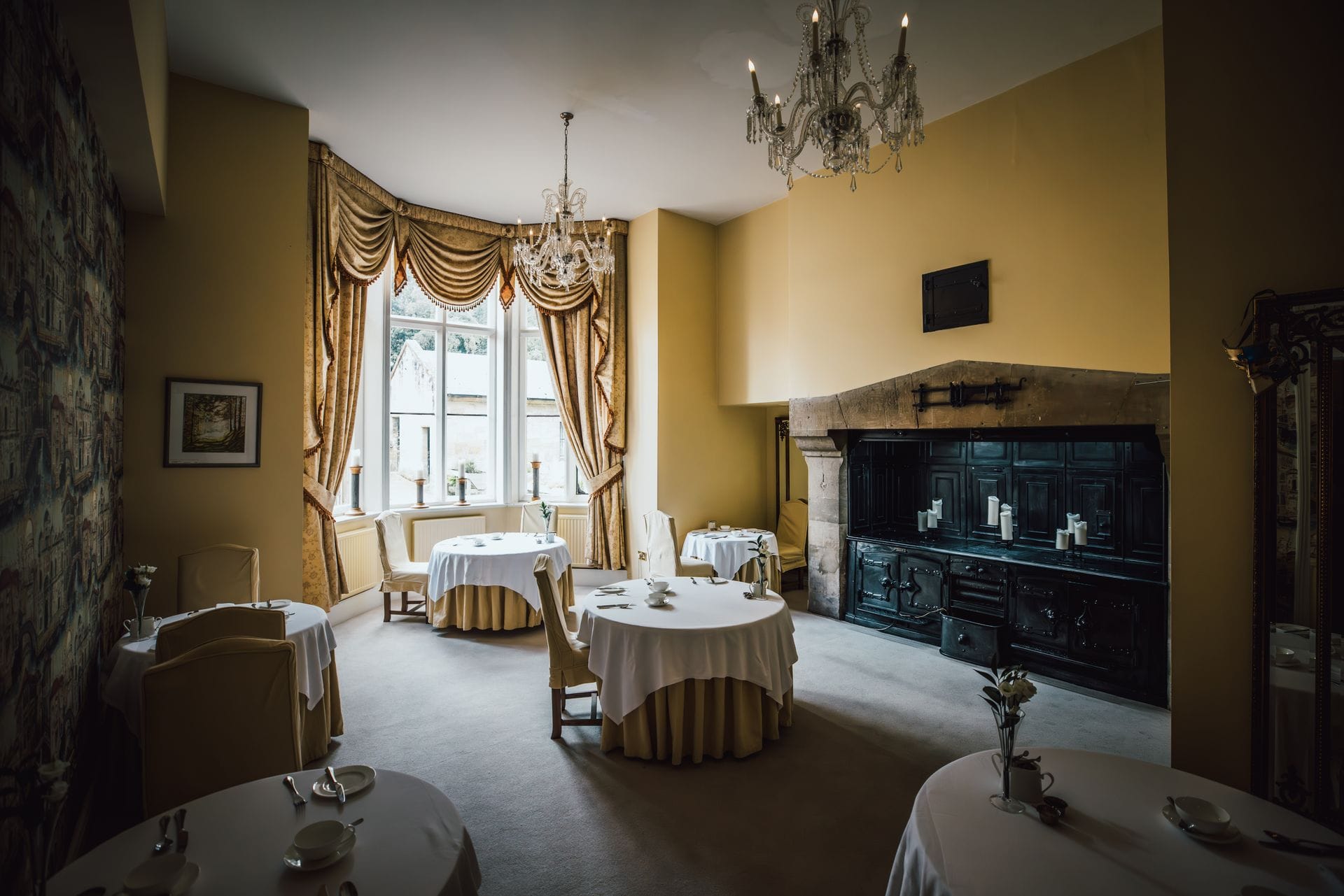 A quiet Welsh boutique hotel dining room set for an intimate dinner service, with a small round table for two positioned by a large sash window, white linen tablecloth, polished silver cutlery, a single flickering candle and glasses of red wine, with soft golden evening light filtering through the window and pale grey painted panelled walls creating a peaceful adults-only fine-dining atmosphere.