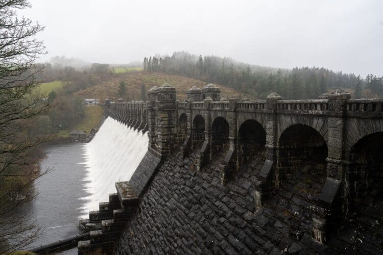 A dramatic view of a massive, historic dark stone dam featuring a series of classic archways along its crest. A powerful, smooth sheet of white water cascades down the sloping face of the structure into the dark river below. The dam is set against a moody, atmospheric backdrop of rolling hills covered in pine forests and autumnal trees, partially shrouded in low-hanging mist under an overcast sky.
