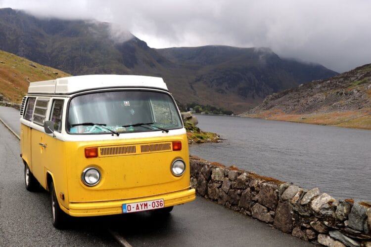 A yellow vintage Wales campervan is parked on a narrow road beside a stone wall, overlooking a lake with misty mountains in the background under a cloudy sky.