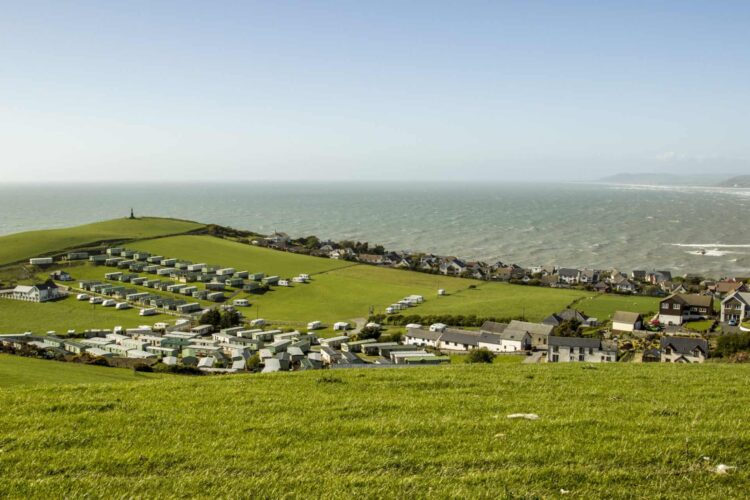 A coastal landscape with green grassy hills, rows of caravans or mobile homes at Brynrodyn Caravan Park in Wales, a cluster of buildings, and the sea in the background under a clear blue sky.