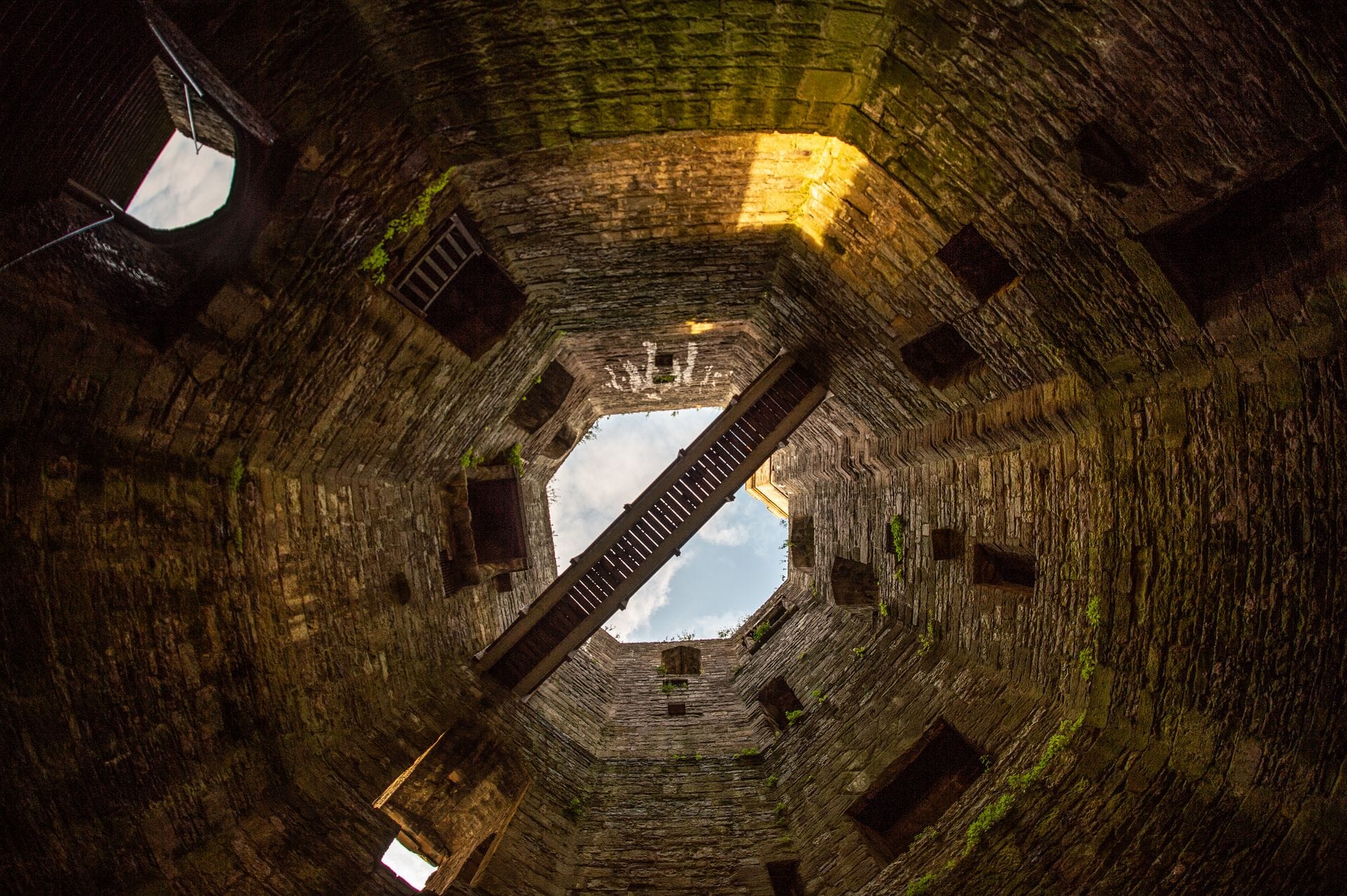 A view looking straight up inside one of Caernarfon Castle's towers, showing wooden internal staircases zigzagging up the stone walls to a small square of blue sky at the top.