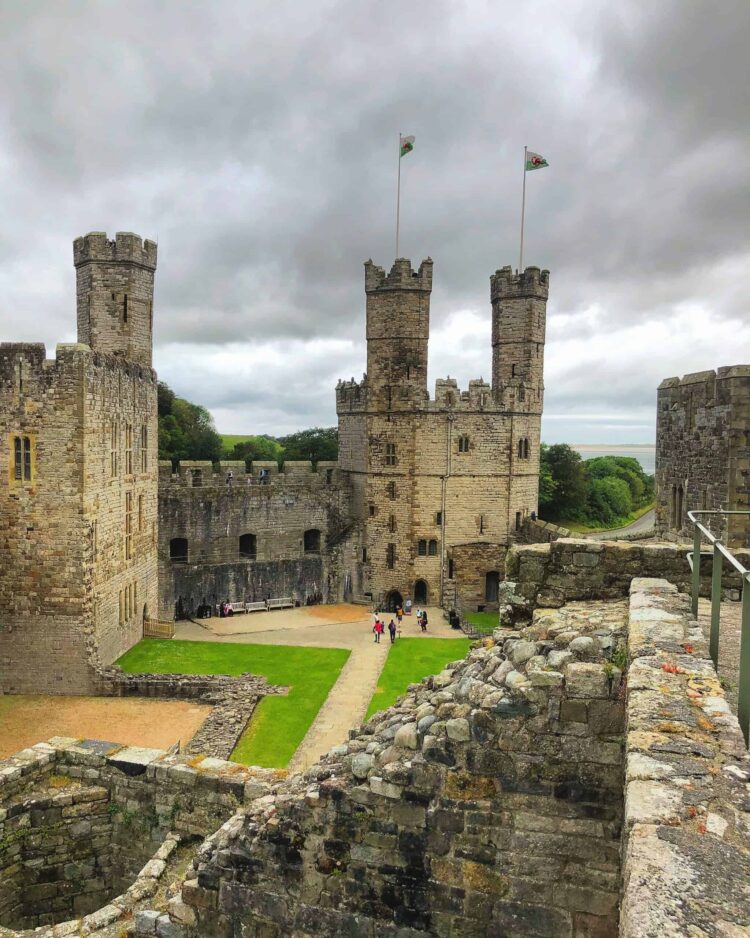 Caernarfon Castle with tall towers and flags sits under a cloudy sky. People are walking in the grassy courtyard surrounded by ancient stone walls. Trees and hills are visible in the background.