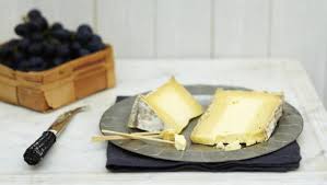 A plate with two wedges of cheese sits on a dark serviette, with cheese knives beside it. In the background, a wooden basket filled with dark grapes is visible—a scene reminiscent of enjoying the national dish of Wales.