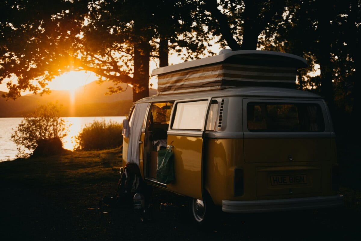 A classic yellow and white campervan parked beneath silhouette trees beside a calm lake at sunset. The pop-top roof is raised and the sliding door is open, with golden evening light reflecting off the water and distant hills in the background.