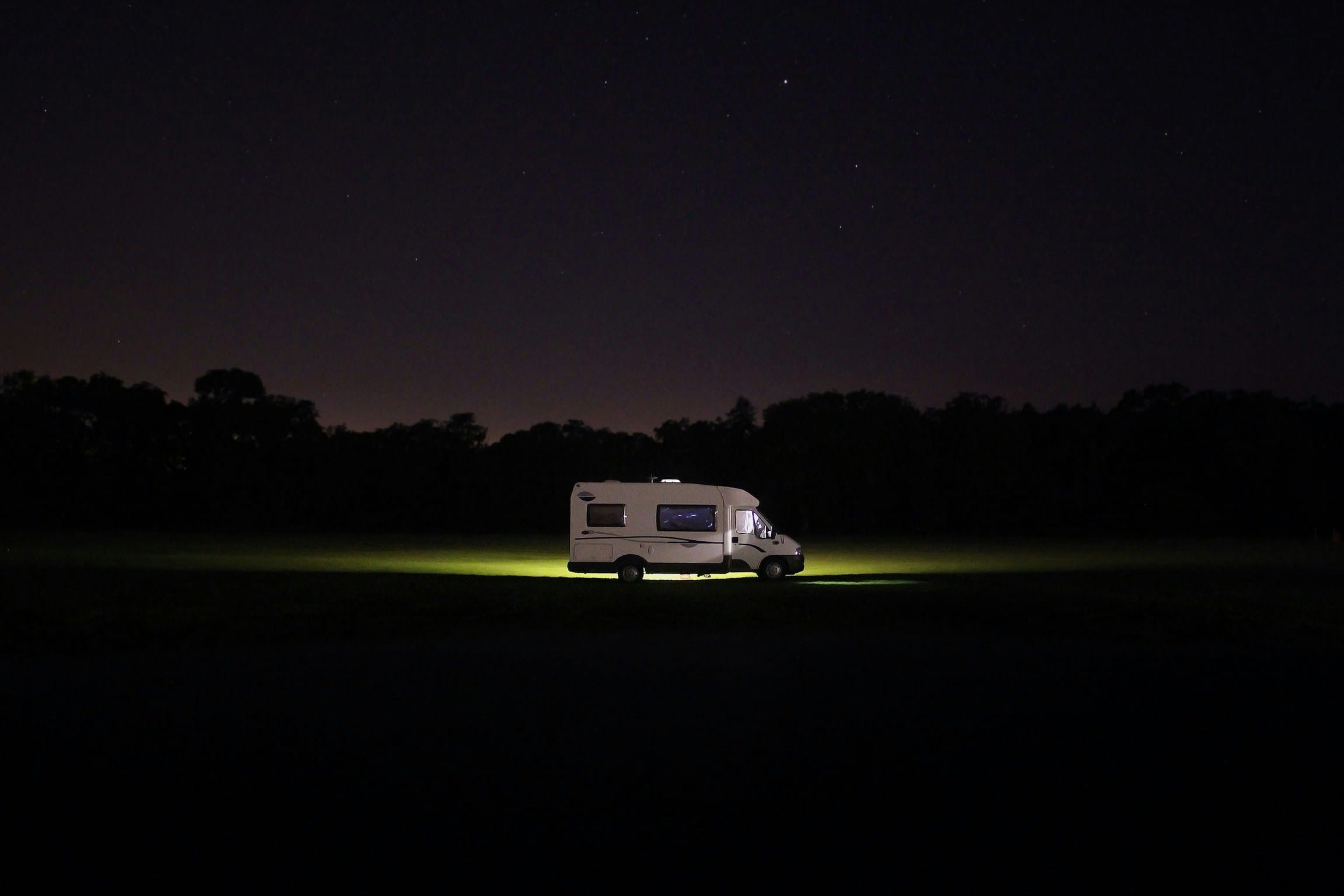 A white motorhome parked in a dark field at night, with bright under-vehicle lights illuminating the grass beneath it against a silhouetted tree line and a starry sky.