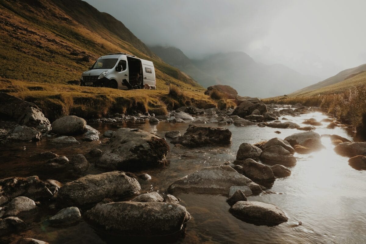 A white campervan parked on a grassy bank next to a rocky, flowing stream in a moody, overcast Welsh mountain valley, highlighting wild North Wales touring sites.