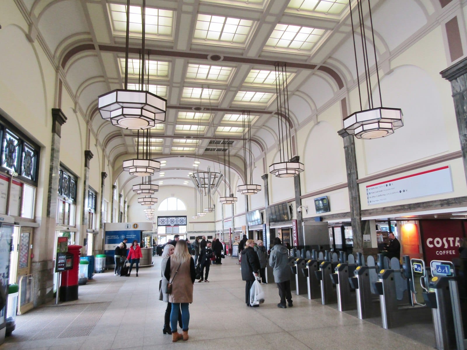 The spacious interior concourse of Cardiff Central railway station, featuring high ceilings with skylighted panels, large hanging Art Deco light fixtures, and ticket barriers on the right. Several passengers are walking through the bright, stone-lined hall, with a Costa Coffee shop visible in the background.