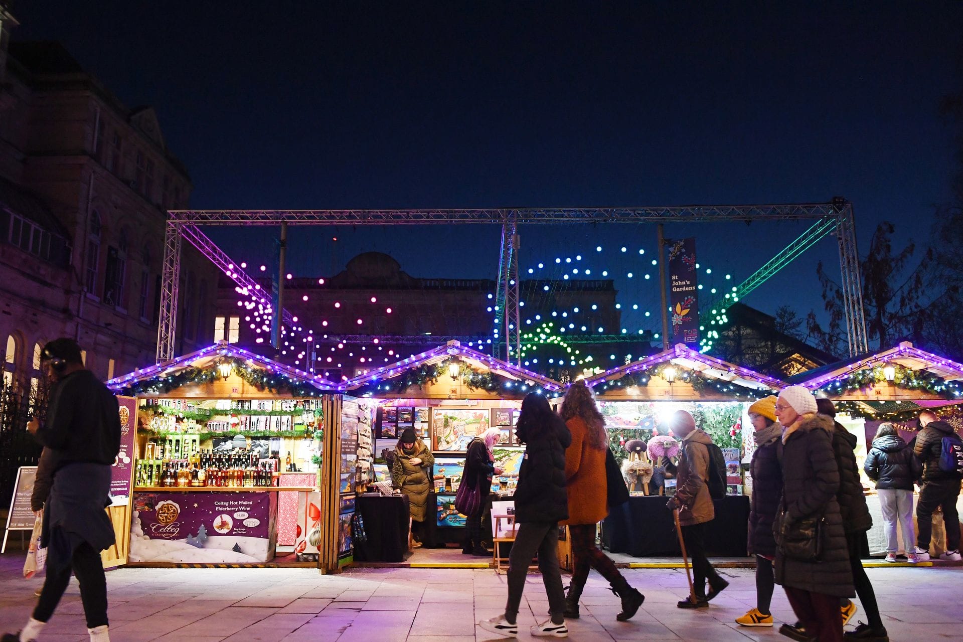 A nighttime view of a bustling Christmas market. Shoppers walk past a row of wooden chalet stalls with warmly lit interiors and peaked roofs, while an overhead display of bright purple and green festive lights illuminates the dark sky above.