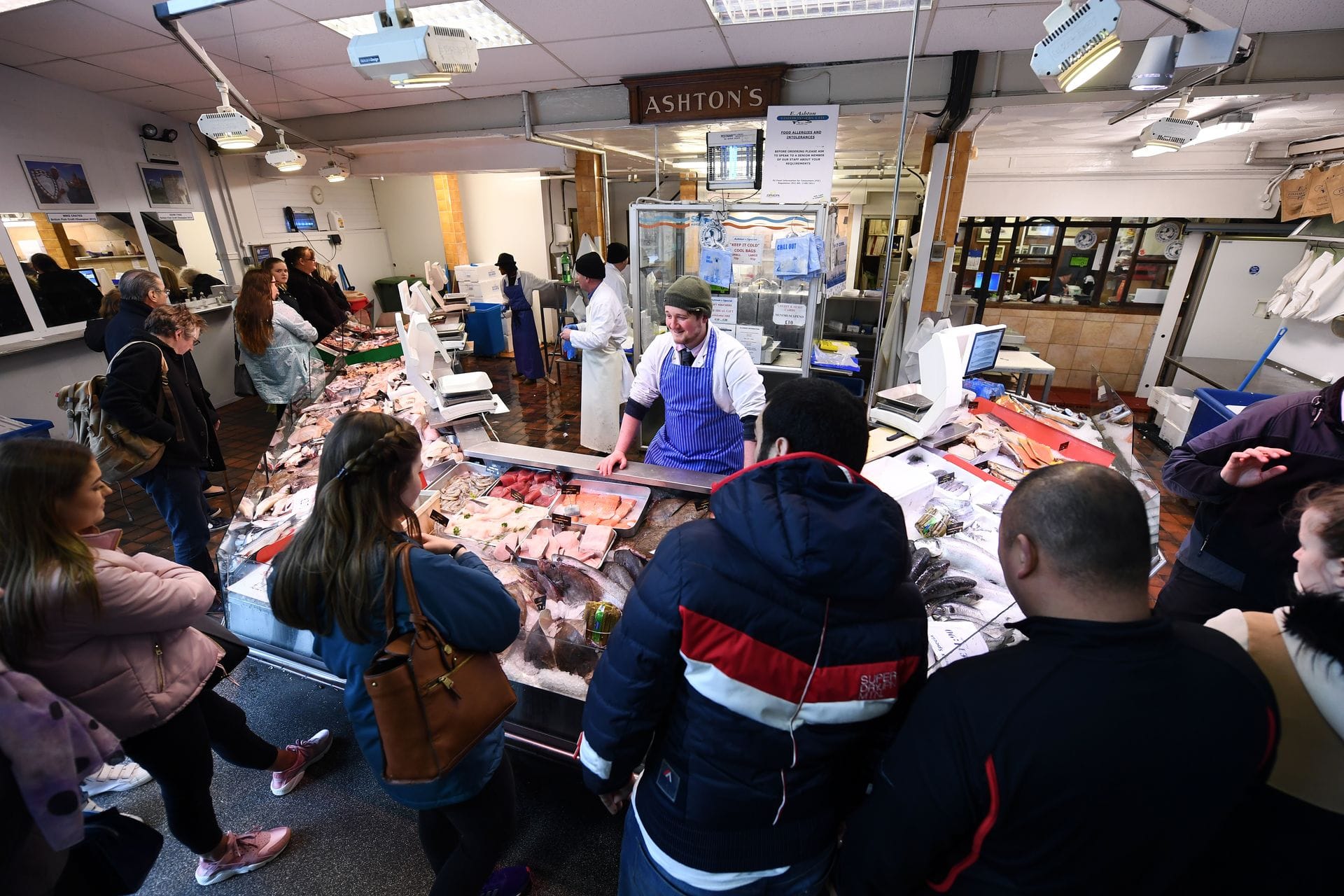 A bustling fishmonger stall named Ashton's inside Cardiff Indoor Market, with a fishmonger in a blue and white striped apron serving customers gathered around a display of fresh seafood on ice.