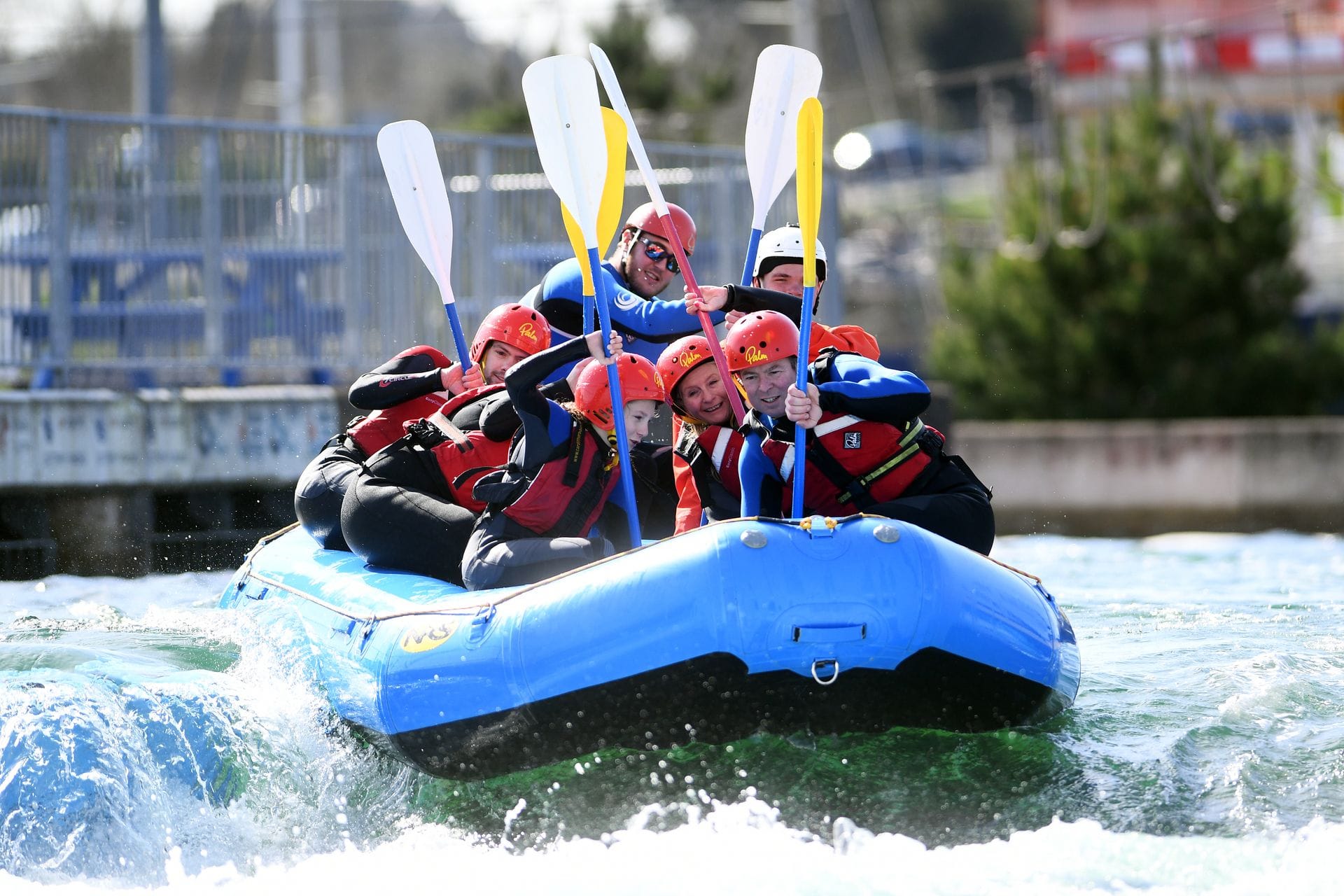 A group of smiling people wearing wetsuits and red helmets raising their paddles in celebration while white water rafting in a bright blue raft at Cardiff International White Water in Cardiff Bay, Wales.