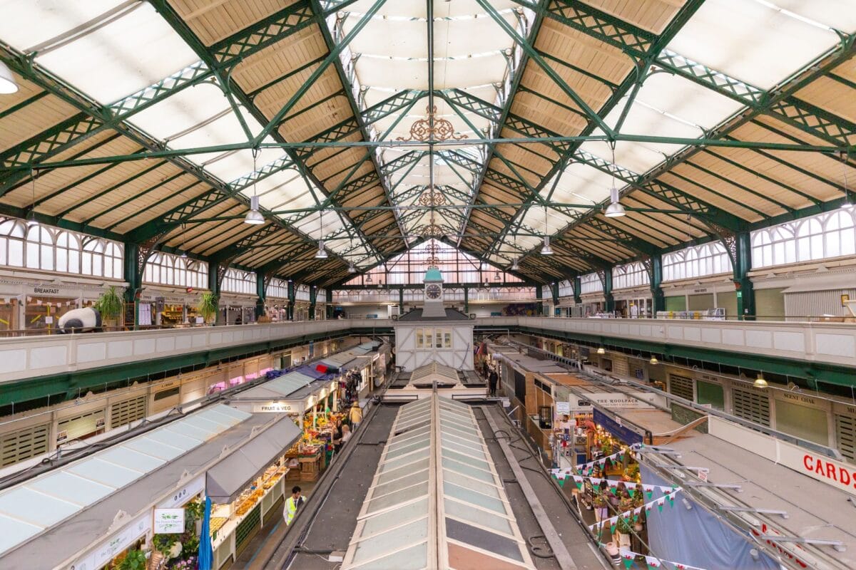 Looking down from the balcony inside Cardiff Central Market. A large Victorian glass and green iron roof covers rows of independent food stalls and shoppers, showcasing the vibrant local culture