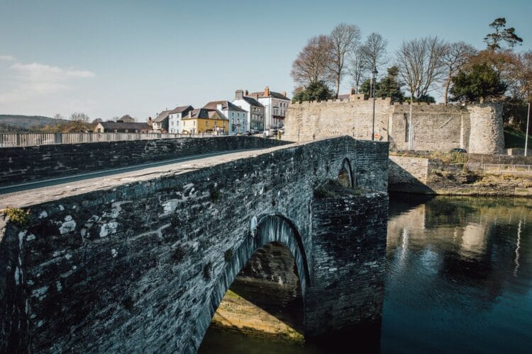 A view of a historic stone arch bridge crossing a calm river, leading toward a town. On the right riverbank, the imposing stone walls and a round tower of Cardigan Castle stand tall, reflecting gently in the water below. In the background, colorful town buildings and bare winter trees sit beneath a clear, bright sky.
