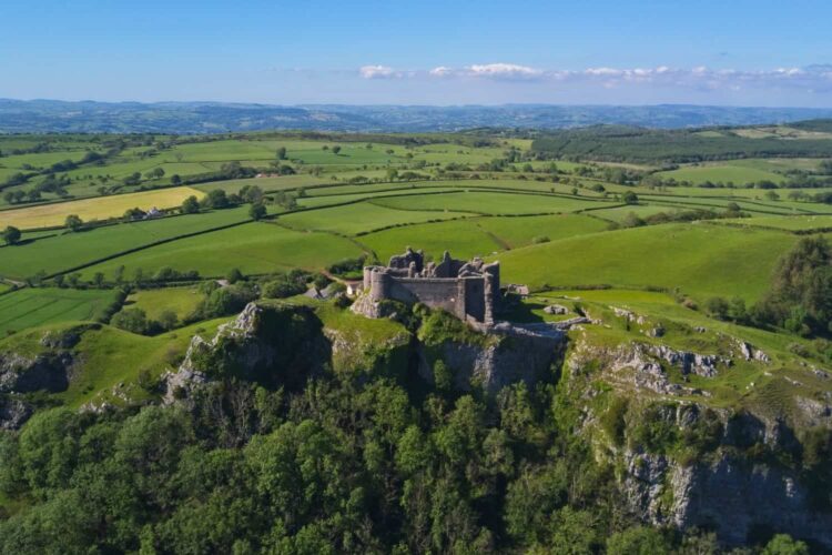 Aerial view of Carreg Cennen Castle ruins perched on a hilltop, surrounded by lush green fields, trees, and rolling countryside under a partly cloudy blue sky.