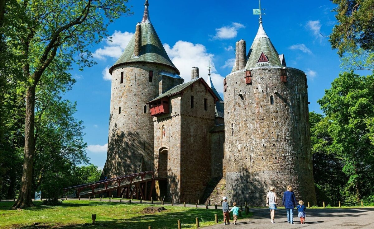 A family of five walking along a paved path toward the massive circular stone towers and pointed conical roofs of Castell Coch near Cardiff, surrounded by bright green summer trees.