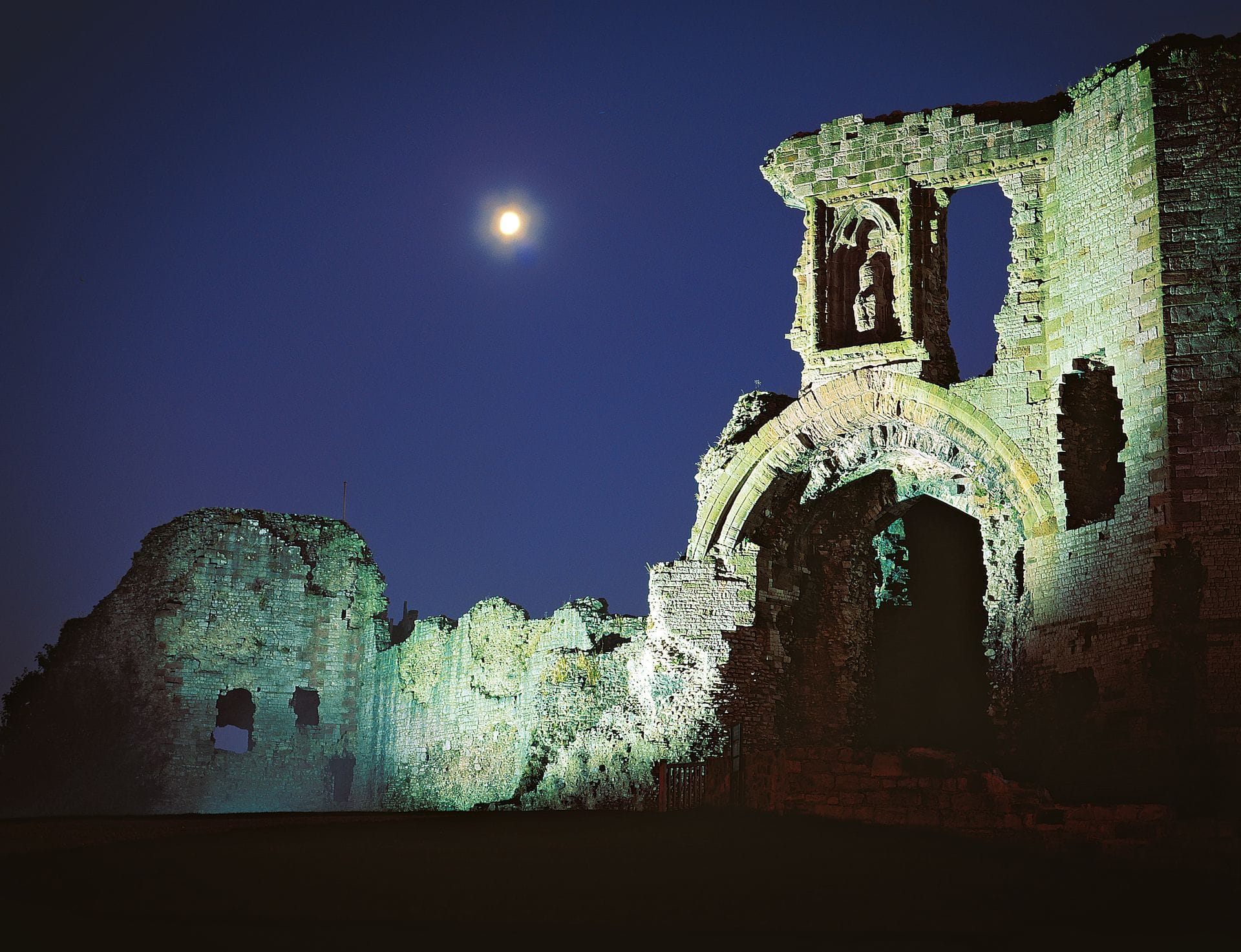 A ruined gothic archway at Denbigh Castle illuminated in dramatic green and white light at night, with a bright full moon visible in the dark sky above.