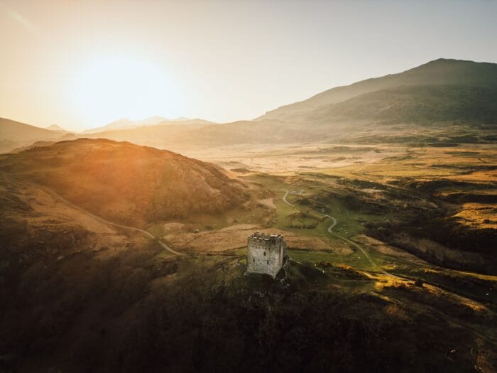 Aerial view of Dolwyddelan Castle, an old stone tower on a grassy hill in Eryri National Park at sunrise. Surrounded by rolling hills and mountains under a hazy sky, winding paths lead through the breathtaking landscape.