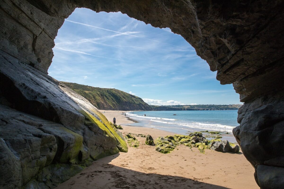 Looking out from inside a rocky sea cave onto the golden sands and green cliffs of Penbryn in Cardigan Bay, highlighting why it is considered one of the best beaches in Wales.