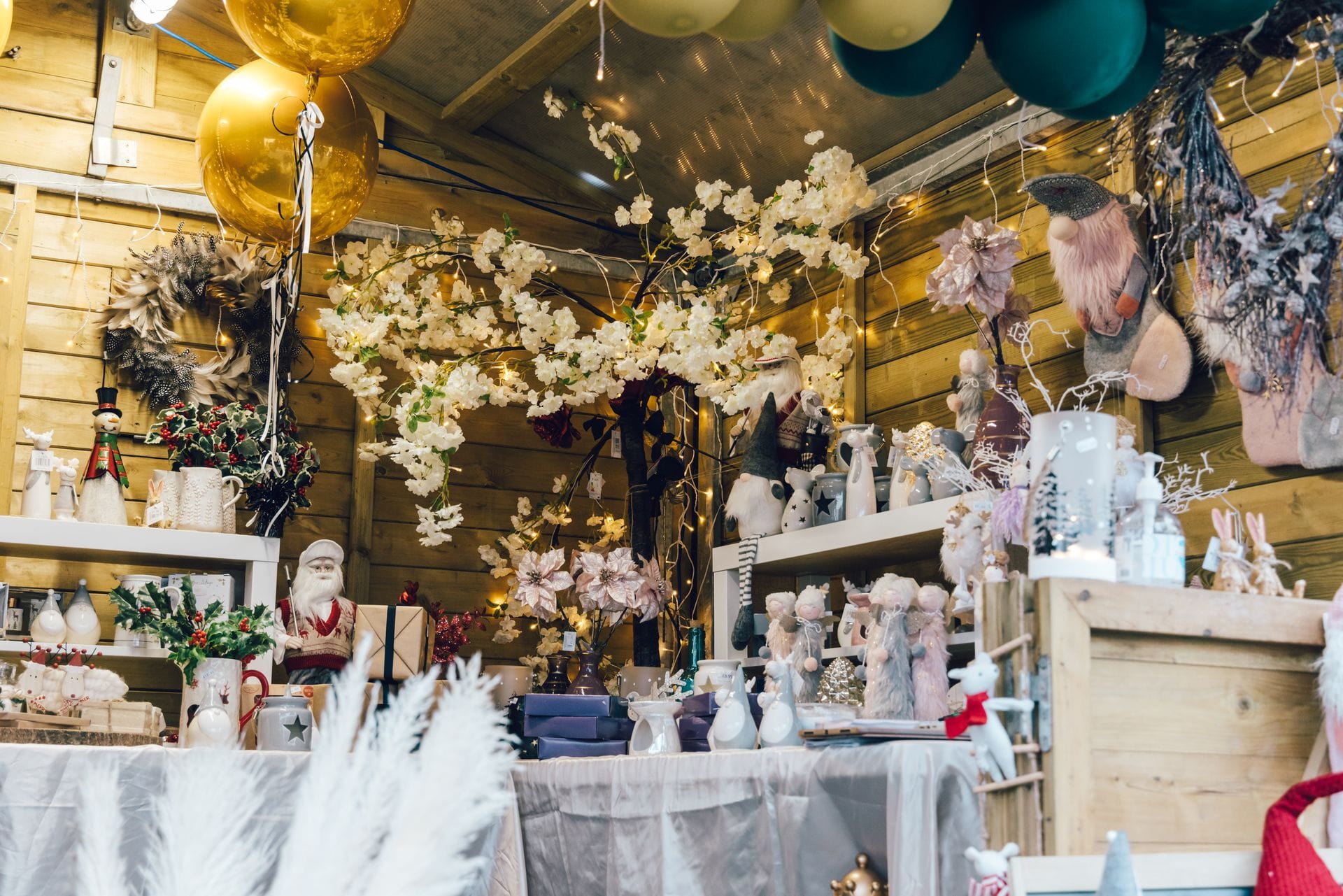A warmly lit wooden market stall filled with festive Christmas decorations. The display includes plush holiday gnomes, white decorative branches, large gold and green baubles hanging from the ceiling, and various small winter figurines arranged carefully on white-draped tables and wooden shelves.