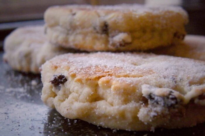 Close-up of sugar-dusted Welsh cakes with visible sultanas, stacked on a dark surface. The warm, homemade feel and tempting texture invite you to try Welsh cakes—one of the must-try things to do in Wales.