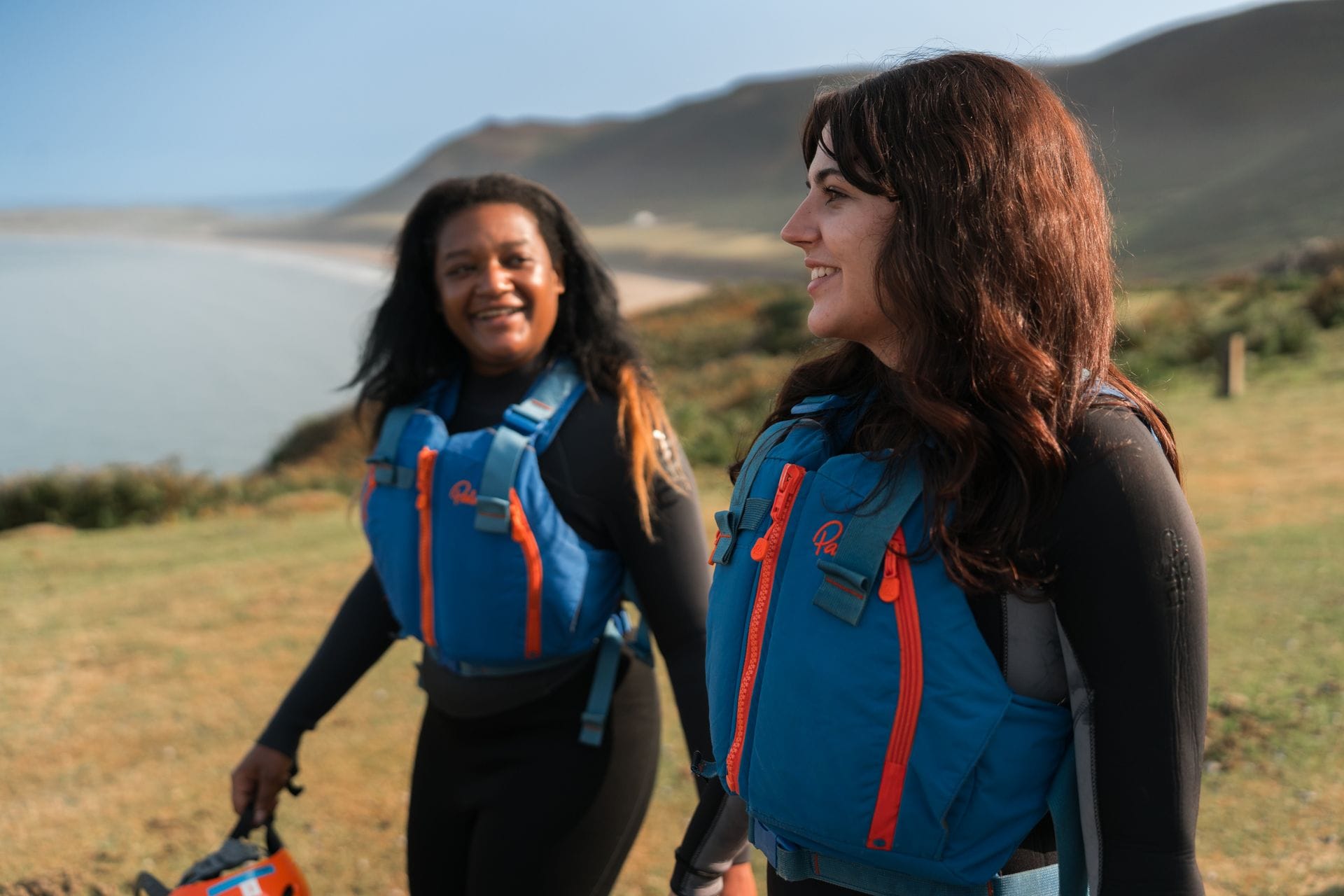 Two smiling women wearing black wetsuits and blue buoyancy aids walking along a grassy coastal clifftop, with a sweeping sandy beach and the ocean in the background.